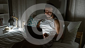 Young arab man using laptop sitting on the bed wearing headphones at bedroom