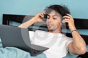 Young arab man using laptop wearing headphones watching film sitting in bed at home. Educational internet website