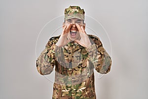 Young arab man wearing camouflage army uniform shouting angry out loud with hands over mouth