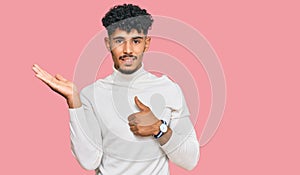 Young arab man wearing casual winter sweater showing palm hand and doing ok gesture with thumbs up, smiling happy and cheerful