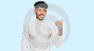 Young arab man wearing casual winter sweater smiling with happy face looking and pointing to the side with thumb up