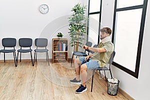 Young arab man wearing neck collar looking watch sitting on chair at hospital waiting room