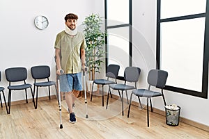 Young arab man wearing neck collar standing using crutches at hospital waiting room