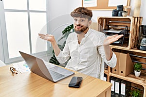 Young arab man working using computer laptop at the office clueless and confused expression with arms and hands raised