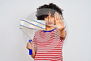 Young arab painter woman with curly hair holding paint roller over isolated white background with open hand doing stop sign with