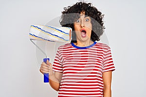 Young arab painter woman with curly hair holding paint roller over isolated white background scared in shock with a surprise face,