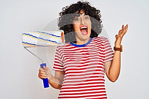 Young arab painter woman with curly hair holding paint roller over isolated white background very happy and excited, winner