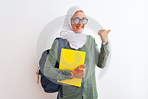 Young Arab student woman wearing hijab and backpack holding a book over isolated background pointing and showing with thumb up to