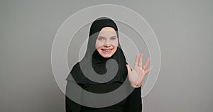 Young arab teenager female muslim student in black hijab waves hello gesture smiling isolated on gray background. Copy