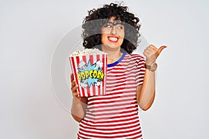 Young arab woman with curly hair holding pack of popcorns over isolated white background pointing and showing with thumb up to the