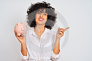 Young arab woman with curly hair holding piggy bank over isolated white background very happy pointing with hand and finger to the