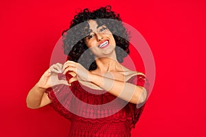 Young arab woman with curly hair wearing casual dress over isolated red background smiling in love showing heart symbol and shape