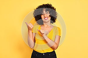 Young arab woman with curly hair wearing t-shirt standing over isolated yellow background Pointing to the back behind with hand