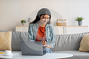 Young Arab woman in headphones using laptop, working or studying online from home, taking notes during business meeting