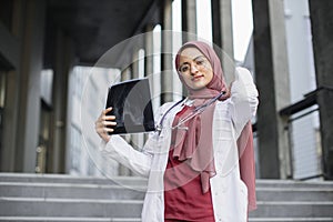 Young Arab woman physician in headscarf holding x-ray, standing outside modern clinic