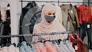 Young arab woman shopper in medical protective mask chooses clothes in clothing store retail sales discount purchase