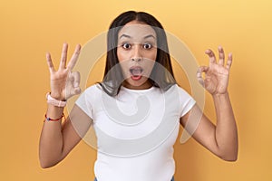 Young arab woman wearing casual white t shirt over yellow background looking surprised and shocked doing ok approval symbol with