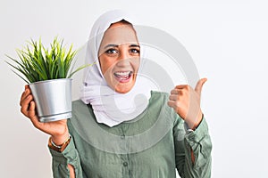 Young Arab woman wearing hijab holding plant pot over isolated background pointing and showing with thumb up to the side with