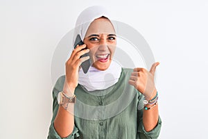 Young Arab woman wearing hijab talking on smartphone over isolated background pointing and showing with thumb up to the side with