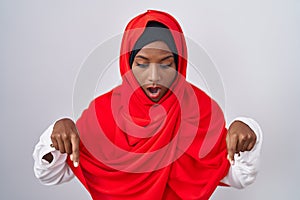 Young arab woman wearing traditional islamic hijab scarf pointing down with fingers showing advertisement, surprised face and open