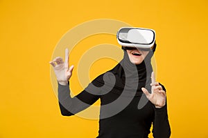 Young arabian muslim woman in hijab black clothes watching in headset of vr virtual reality isolated on yellow wall