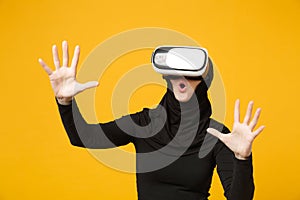 Young arabian muslim woman in hijab black clothes watching in headset of vr virtual reality isolated on yellow wall
