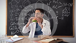 Young Arabic chemistry teacher holding a piece of hamburger. Student eating fast food in the office. Burger is not