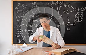 Young Arabic chemistry teacher holding a piece of hamburger. Student eating fast food in the office. Burger is not