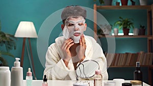 young Arabic man applies moisturizing sheet mask on face dressed in white bathrobe. Male daily beauty routine