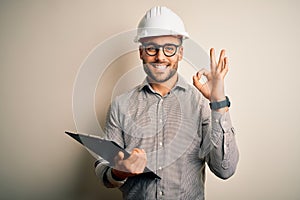 Young architect inspector man wearing builder safety helmet and using clipboard doing ok sign with fingers, excellent symbol