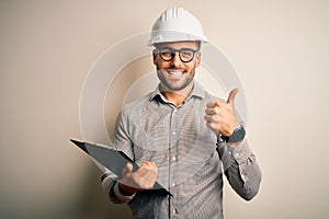 Young architect inspector man wearing builder safety helmet and using clipboard happy with big smile doing ok sign, thumb up with