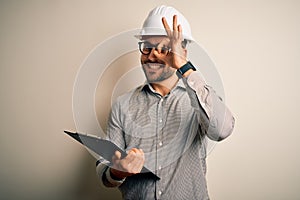 Young architect inspector man wearing builder safety helmet and using clipboard with happy face smiling doing ok sign with hand on