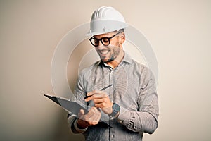 Young architect inspector man wearing builder safety helmet and using clipboard with a happy face standing and smiling with a