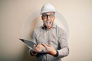 Young architect inspector man wearing builder safety helmet and using clipboard very happy pointing with hand and finger