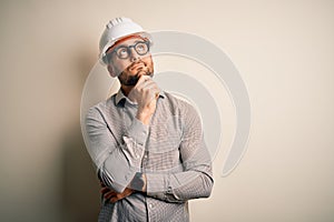 Young architect man wearing builder safety helmet over isolated background with hand on chin thinking about question, pensive