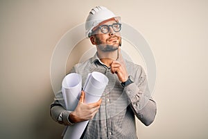 Young architect man wearing contractor helmet holding project paper plan over isolated background serious face thinking about