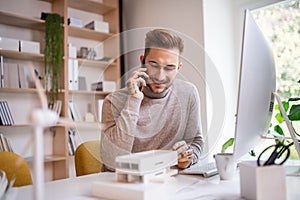 Young architect sitting at the desk indoors in office, using smartphone.