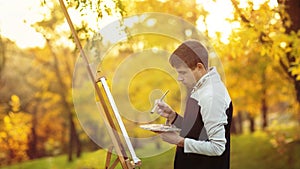 Young artist drawing a picture on canvas on an easel in nature, a man with a brush and a painter gamut among autumn trees, a
