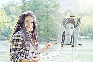 Young artist girl painting a self portrait in a park near lake - Painter woman with dreadlocks hairstyle working on her art