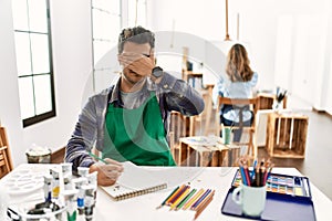 Young artist man at art studio covering eyes with hand, looking serious and sad