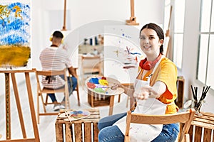 Young artist woman painting on canvas at art studio smiling cheerful offering palm hand giving assistance and acceptance