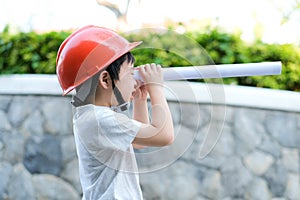 Young Asia boy in gray T shirt wearing a orange safety helmet and holding blueprints in the garden at home