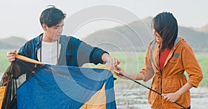 Young asia campers couple setting up the tent camping gear outdoor near seaside. Male and female travel having fun on a summer day