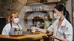 Young Asia female barista wear face mask serving take away hot coffee paper cup to consumer at cafe. Owner small business,