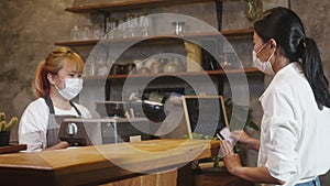 Young Asia female barista wear face mask serving take away hot coffee paper cup to consumer and using credit card for payment at