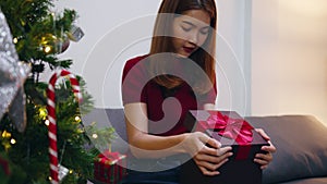 Young Asia female having fun opening X`Mas present box near Christmas tree decorated with ornaments in living room at home. Merry