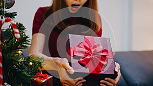 Young Asia female having fun opening X`Mas present box near Christmas tree decorated with ornaments in living room at home. Merry