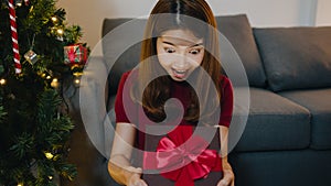 Young Asia female having fun opening X`Mas present box near Christmas tree decorated with ornaments in living room at home. Merry