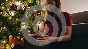 Young Asia female having fun opening X`Mas present box near Christmas tree decorated with ornaments in living room at home. Merry