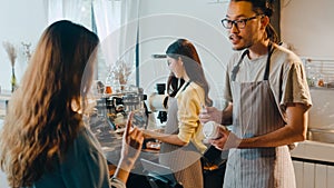 Young Asia male barista waiter taking order from customer standing behind bar counter while talking with customer making note on